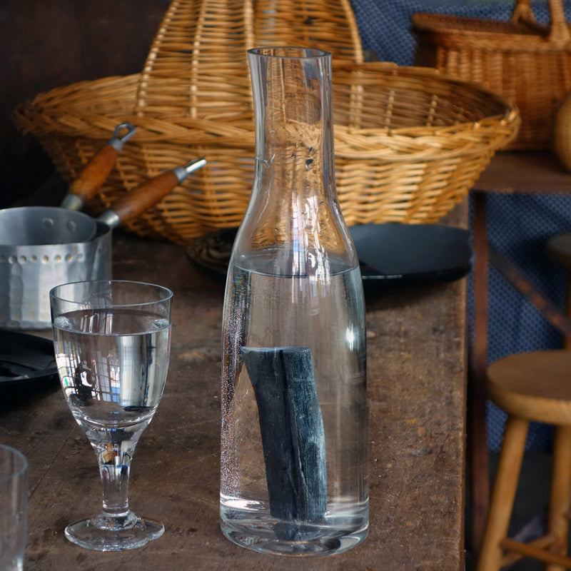carafe en verre artisanale de forme irrégulière avec un morceau de charbon devant des paniers en osier - tsé tsé